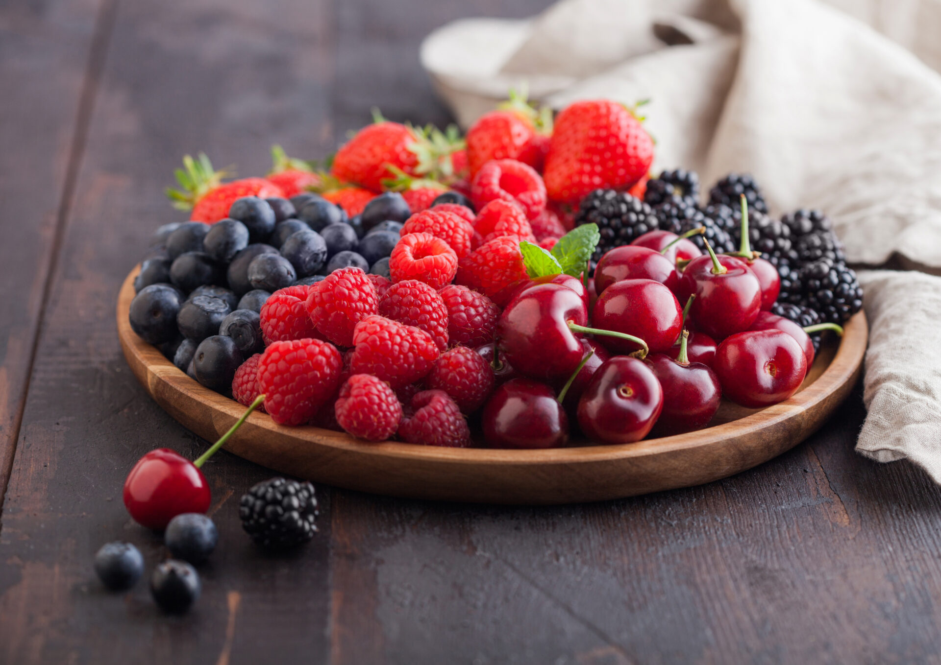 Fresh organic summer berries mix in round wooden tray on dark wooden table background. Raspberries, strawberries, blueberries, blackberries and cherries with linen kitchen towel. Top view
