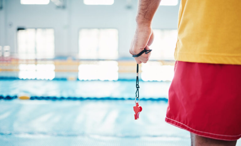 Hands, lifeguard and whistle by swimming pool for water safety, security or ready for rescue indoors. Hand of expert swimmer holding signal tool for warning, safe swim or responsibility for awareness